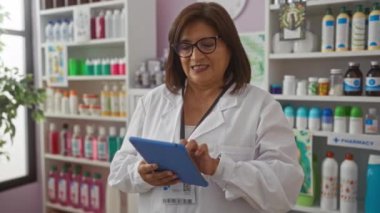 Etrafı çeşitli tıbbi ve kozmetik ürünlerle çevrili iyi stoklanmış bir eczanede beyaz laboratuvar önlüğü ve gözlük takan İspanyol kadın eczacı..