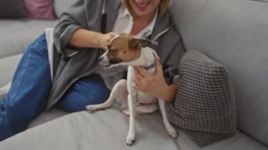Orta yaşlı sarışın bir kadın, modern oturma odasındaki rahat gri bir kanepede otururken küçük bir köpeği nazikçe fırçalıyor..
