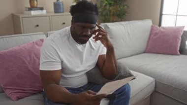 Sakallı genç bir adam, evinin oturma odasındaki bir kanepede oturur ve rahat bir evde özenle bir belge okur..