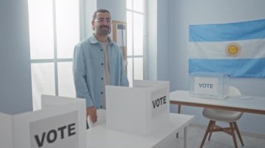 Arjantinli bir seçim okulunda oy veren genç bir adam, oy verme kabinleri ve bir Arjantin bayrağıyla çevrelenmiş bir jest yapıyor.