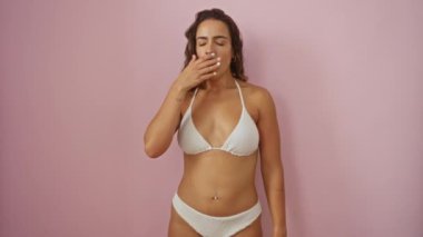 Esneyen, bikinili genç bir kadın izole edilmiş pembe arka planda oturuyor. Uykusuzluk yüzünden ağzını kapatmaktan sıkılmış ve yorgun görünüyor.