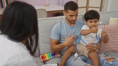 Baba ve oğul steteskopla oynarken kadınlar evlerinde oturup aile içi etkileşimlerini izliyorlar.