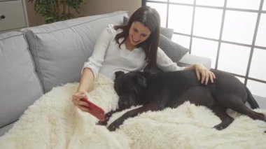 Kentin iç kısımlarında gri bir koltukta uzanmış İspanyol kadın siyah labrador köpeğiyle selfie çekerken gülümsüyor..