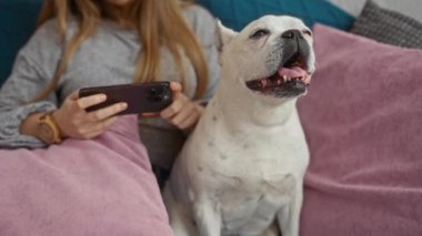 Evde telefonuyla rahatlayan genç bir kadın ve rahat bir oturma odasındaki kanepede köpek yavrusu..