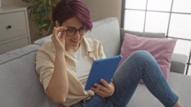 Kısa saçlı ve gözlüklü genç bir kadın evindeki oturma odasında kanepede dinlenirken tablet kullanıyor..