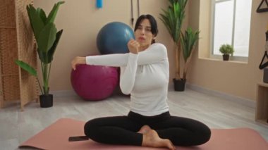 Spor salonunda egzersiz yapan bir kadın yoga minderinin üzerinde egzersiz yapıyor ve arka planda bitkiler var.