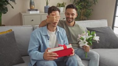 Oturma odalarında aşkı kutlayan iki adam, çiçekler ve bir hediyeyle, beraberlik, neşe ve aile ve evin sıcaklığını sergiliyorlar.