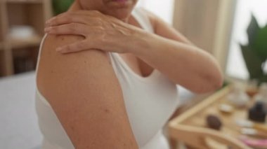 Yaşlı bir kadın spa ortamında omzunu içeride ovalıyor ve yanında sağlık ürünleri var.