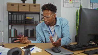 Ofis ortamındaki genç, Afro-Amerikalı bir adam çeşitli ofis malzemeleriyle dolu bir masada otururken düşünceli ve şaşkın görünüyor..