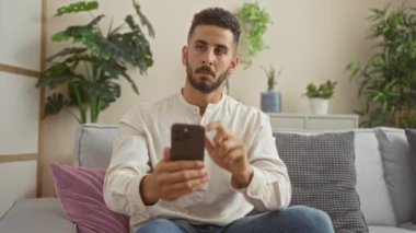 Yakışıklı, İspanyol asıllı genç bir adam akıllı bir telefon tutuyor. Düşünceli bir şekilde oturma odasındaki kanepede oturuyor. İçinde çeşitli bitkiler var..