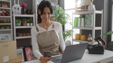 Bir kadın akıllı telefonunu ve laptopunu arka plandaki raflarda düzgünce yerleştirilmiş eşyalarla kontrol ediyor.