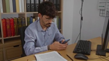 Ofisteki sakallı genç adam arka planda kitap ve dosyalarla dolu bir masada oturmuş gözlüklü ve çizgili gömlekli akıllı telefon kullanıyor.