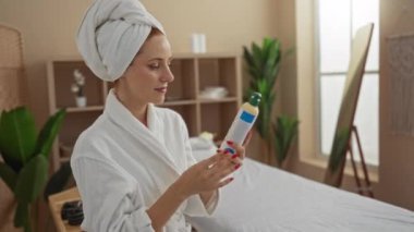 Bornoz ve havlu giymiş bir kadın spa odasında bitki ve spa yataklı bir şişe tutuyor.