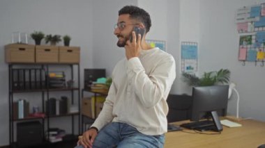 Ofisteki bir telefon görüşmesinde raflarda ve arka planda bitkilerle oturan İspanyol genç bir adam..