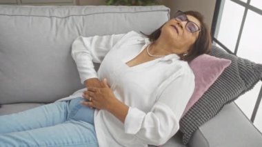 Kısa saçlı, beyaz gömlekli ve kot pantolonlu yaşlı bir kadın evde bir kanepede uzanmış, gözlük takıyor ve kolye ve küpe takıyor..