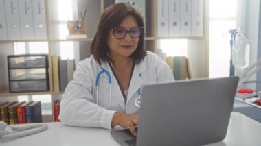 Klinikteki yaşlı bir İspanyol kadın doktor dizüstü bilgisayar üzerinde çalışıyor ve gözlüklerini çıkarıyor. Üzerinde steteskop ile laboratuvar önlüğü var..