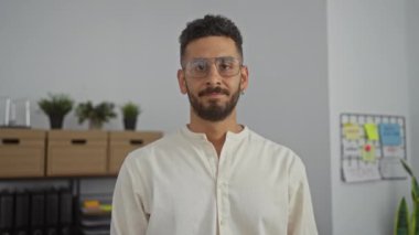 Beyaz tişörtlü çekici bir genç adam arka planda çiçekleri ve beyaz tahtası olan bir ofiste güvenle dikiliyor..