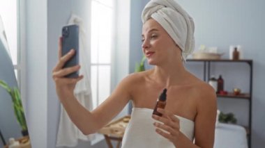 Kafasına havlu sarılmış genç bir kadın, arka planda raflar olan kaplıca benzeri bir odada elinde kozmetik bir şişeyle video görüşmesi yapıyor.