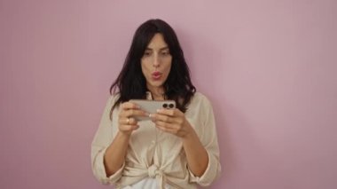 İzole edilmiş pembe arka plan üzerinde akıllı telefon kullanan kadın ekrana odaklanmış, modern günlük kıyafetlerle mutlu ve şaşırmış görünüyor.