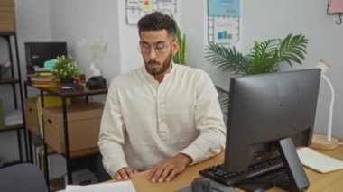 Ofiste çalışan, gözlük takan, bilgisayar ve evrak işleriyle uğraşan, bitkilerle ve ofis malzemeleriyle çevrili, görevlerine odaklanmış,.