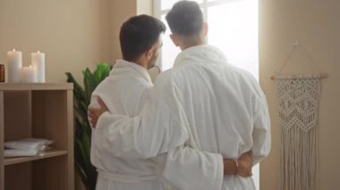 Beyaz cüppeli adamlar, mumlar, bitkiler ve rüya avcılarıyla dolu sakin bir spa odasında kucaklaşıyorlar. Kapalı bir ortamda sevgi ve rahatlamayı yansıtıyorlar.