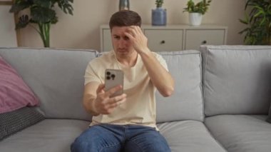 Genç, yakışıklı bir adam evinde bir kanepede oturuyor, akıllı telefonuna bakarken saçını tarıyor..