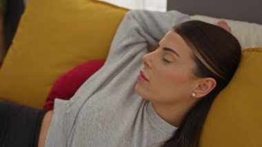 Esmer saçlı güzel bir kadın evdeki rahat bir yatak odasında huzur içinde uyuyor, sarı bir yastığa başını yaslıyor ve rahat gri bir bluz giyiyor..