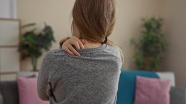 İçeride sarı saçlı bir kadın, arkadan görülmüş, gri bir süveter giyiyor ve elleri omuzlarında, rahat bir oturma odasında pembe ve mavi yastıklı ve yeşil bitkilerli..