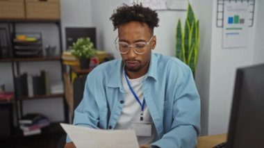 Sakallı ve gözlüklü genç bir siyah adam arka planda bitki ve rafların olduğu bir ofis ortamında dikkatle belgeleri okuyor..