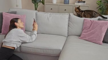 Rahat bir oturma odasında genç bir kadın kanepede uzanan kedisiyle iletişim kurmak için akıllı telefonunu kullanıyor..