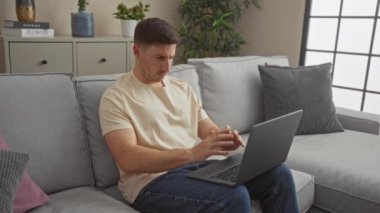 Genç bir adam, rahat bir oturma odasında dizüstü bilgisayarla otururken, etrafı bitkiler ve yastıklarla çevrili bir şekilde, evinde dinlenme anı yakalıyor..