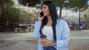 Açık hava parkında elinde kahve fincanı tutarken akıllı telefondan konuşan çekici, genç esmer kadın..
