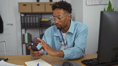 Sakallı ve gözlüklü Afro-Amerikalı genç bir adam, düzenli raflar ve bilgisayarla çevriliyken telefonunu kullanarak bir ofiste çalışıyor..