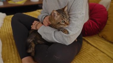 Yatak odasındaki rahat sarı bir yatakta oturan bir kadın, kollarını çizgili bir kediye dolamış, rahatlatıcı bir iç mekan sergiliyor..