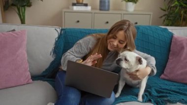 Dizüstü bilgisayarı ve köpek evcil köpeği olan genç sarışın kadın oturma odasının iç kısmında kanepede oturuyor..