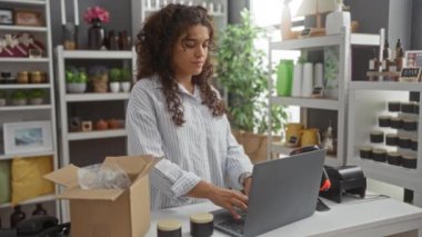 İç mekan dekorasyonunda çalışan genç bir İspanyol kadın önündeki dizüstü bilgisayarla paketleri taşıyordu ve rafların arkasındaki ürünleri düzgünce düzenliyordu..