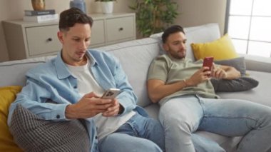 Modern oturma odasındaki koltukta akıllı telefon kullanan erkekler, evde teknolojiden keyif alan eşcinsel çiftleri ön plana çıkarıyorlar.