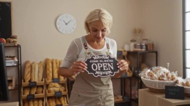 Elinde ekmek rafları ve glütensiz ürünlerle fırında açık bir tabela tutan kadın, gülümsüyor, önlük giymiş, saatin yanında duruyor, yüzünde hoş bir jest gösteriyor.