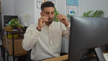 Gözlük takan genç bir İspanyol adam modern bir ofiste bilgisayarının başında çalışıyor. Arka planda iç mekanlar ve organize raflar var..