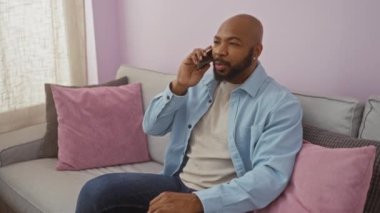 Evdeki bir kanepede oturan genç adam pembe yastıkları ve yumuşak bir ambiyansı olan oturma odasında telefonla konuşuyor.
