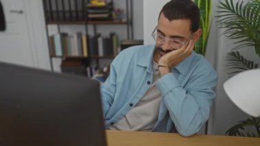Bıyıklı ve gözlüklü genç adam bilgisayara dikkatlice bakıyor. Etrafı kitaplarla ve ev bitkileriyle çevrili.