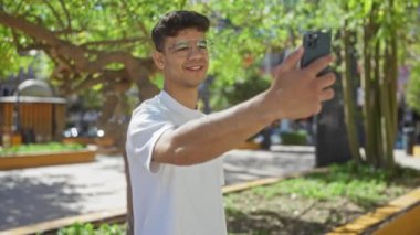 Genç İspanyol bir adam parkın şehir merkezinde akıllı telefonuyla selfie çekiyor güneşli bir günde mutlu bir şekilde gülümsüyor..