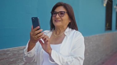 Orta yaşlı, kısa saçlı, beyaz bluz ve gözlük takan bir İspanyol kadın akıllı telefonunu şehir caddesinde kullanıyor..