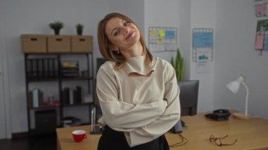 Sarışın, genç bir kadın modern bir ofiste sırayla gülümsüyor. Çalışma masaları ve rafları gibi ofis dekorlarıyla çevrili..