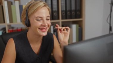 Kulaklık takan genç bir kadın arka planda klasörlerle dolu rafları olan bir ofiste konuşuyor ve gülümsüyor.