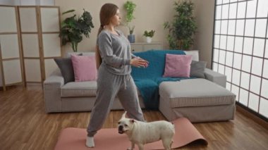 Genç bir kadın, rahat bir oturma odasında köpeğiyle egzersiz yapıyor. Çiçekler, kanepe ve yoga minderiyle birlikte..