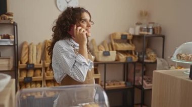 Ekmek ve pastalarla çevrili fırında telefonla konuşan genç bir kadın.