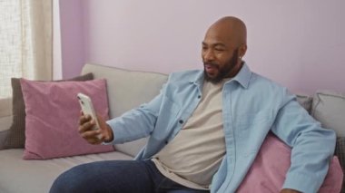 Genç adam rahat bir oturma odasında akıllı telefonuyla video görüşmesi yapıyor, pembe yastıklı bir koltukta oturuyor, mavi gömlek ve kot pantolon giyiyor, anın tadını çıkarıyor.