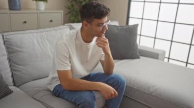 Genç İspanyol bir adam modern bir oturma odasında gri bir kanepede oturuyor. Gözlük, beyaz bir tişört ve kot pantolon giyiyor. Düşünceli bir şekilde çenesini elinin üzerine koyuyor..