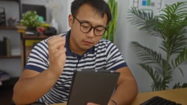 Ofisteki genç Çinli bir adam, bir tablette kitap okuyor. Stresli görünüyor. Etrafı kapalı bitkilerle ve kitaplıklarla çevrili..
